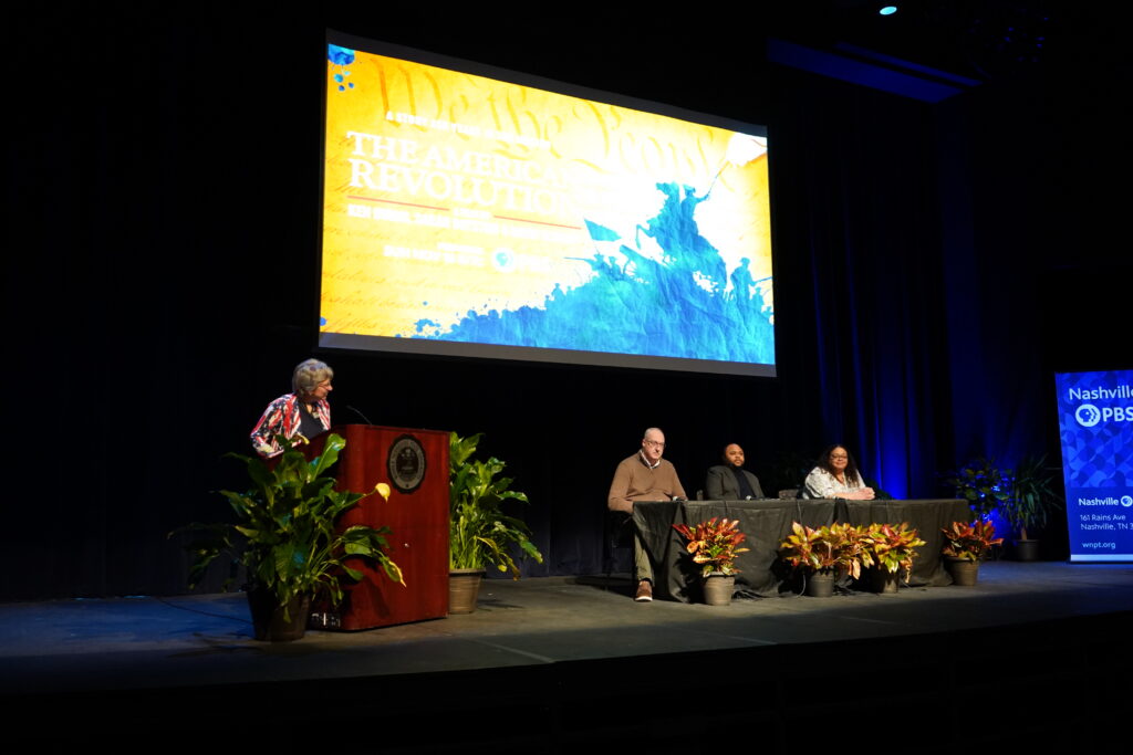 A woman stands at a podium on a stage, three people seated at a table on the stage